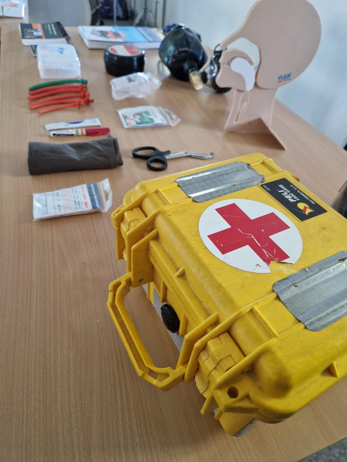 Gelber Notfallkoffer mit rotem Kreuz, dahinter Atemwegshilfen und ein Kopfmodell