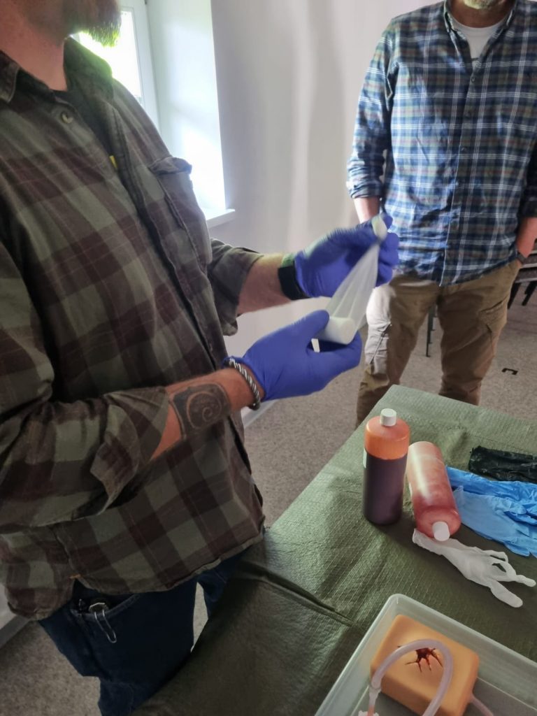 Teilnehmer mit Einmalhandschuhen bereitet Verbandmaterial vor, auf dem Tisch Kunstblut und ein Wundmodell